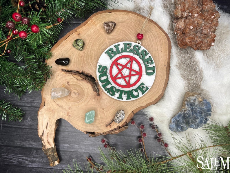 May include: A white wooden ornament with the words "Blessed Solstice" written in green letters around a red pentagram. The ornament is hanging from a wooden slice with various crystals and stones.