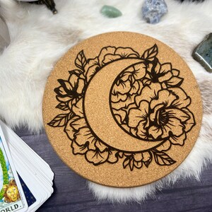 May include: Round cork coaster with a crescent moon and floral design etched in black. The coaster is surrounded by a white fur-like material, with a tarot card deck visible on the left side. Crystals are scattered around the coaster.