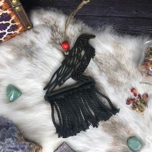 May include: Black wooden raven wall hanging with a macrame fringe, accented by a red bead. The raven is perched on a wooden base. The background includes a white fur-like surface and various crystals.