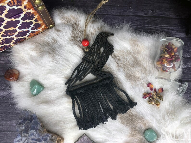 May include: Black wooden raven wall hanging with a macrame fringe, accented by a red bead. The raven is perched on a wooden base. The background includes a white fur-like surface and various crystals.