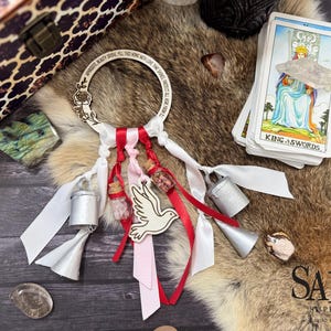 May include: A wooden dream catcher with a rose design and the inscription "APPRECIATE BEAUTY DRIVE ALL THIS HOME WITH LOVE THAT SHINES PROTECT ALL HERE FROM EVIL". It has white ribbons, red and pink ribbons, small glass bottles, and a wooden dove. A deck of tarot cards is also visible.