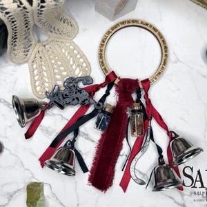 May include: A wooden ring with text, adorned with silver bells, red and black ribbons, small glass bottles, and a decorative metal piece. The design includes a cream-colored lace and various gemstones. The text on the ring reads: "RING GO THE BELLS, EVIL SPIRITS THEY DO REPEL, ALLOW TO PASS THOSE WHO MEAN NO HARM, AS I WILL IT. IT".