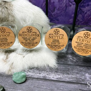 May include: Four glass jars with wooden lids featuring pentagram symbols and engraved text. The jars are labeled with the names of herbs: Hyssop, Red Clover, Nettle, and Vervain. Each label lists the herb's magical properties.