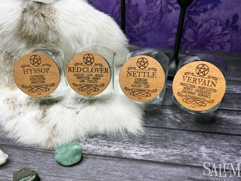 May include: Four glass jars with wooden lids featuring pentagram symbols and engraved text. The jars are labeled with the names of herbs: Hyssop, Red Clover, Nettle, and Vervain. Each label lists the herb's magical properties.