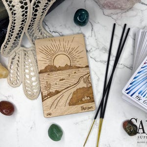 May include: A wooden tarot card with a desert landscape design and the words "The Sun". Incense sticks, crystals, and a deck of tarot cards with the words "the lovers" are also visible. The Salem Supply Co. logo is in the bottom right corner.