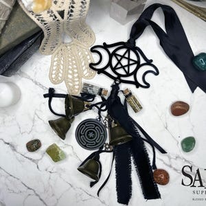 May include: An assortment of mystical items, including bronze bells, a black pentacle pendant, small glass vials, and various gemstones. A white candle and a stack of books are also visible. The items are arranged on a marble surface.