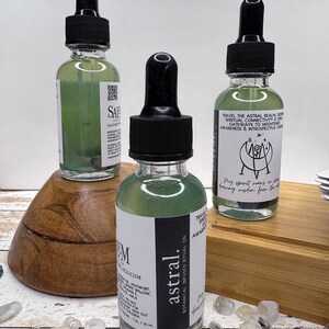 May include: Three glass bottles with dropper tops filled with a green liquid. The bottles are labelled "astral" and "TRAVEL THE ASTRAL REALM, DEEPEN SPIRITUAL CONNECTIVITY & OPEN GATEWAYS TO HEIGHTENED AWARENESS & INTROSPECTIVE VISION". The bottles are sitting on a wooden surface with white and blue stones.