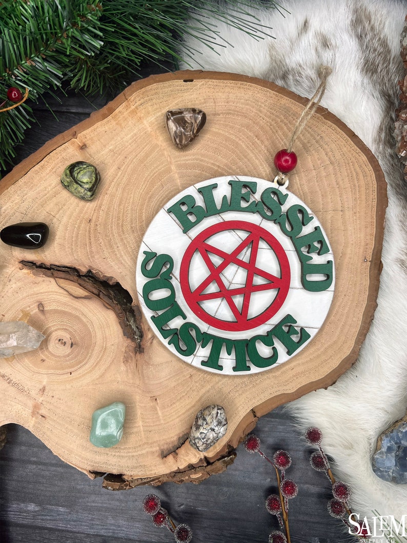 May include: A white wooden ornament with the words 'Blessed Solstice' written in green and red paint. A red five-pointed star is in the center of the ornament.