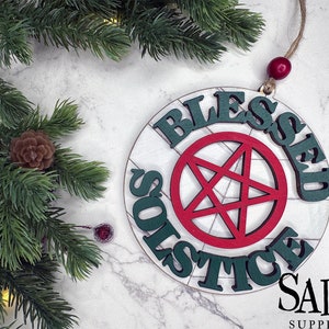 May include: A round wooden ornament with the words "Blessed Solstice" written in green around a red pentagram. The ornament is hanging from a string with a red bead. The background is a white marble surface.