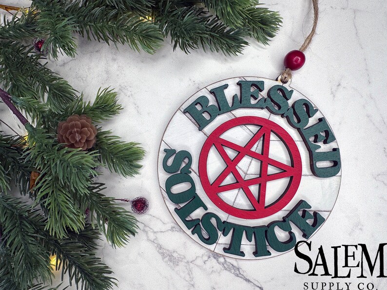 May include: A round wooden ornament with the words "Blessed Solstice" written in green around a red pentagram. The ornament is hanging from a string with a red bead. The background is a white marble surface.