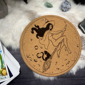May include: A cork coaster with a black and white illustration of a witch flying on a broomstick with a black cat. The witch is wearing a hat and a cape. The background is a cork surface with a pattern of small circles.