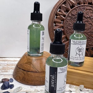 May include: Three glass bottles of green liquid with black dropper tops. The bottles are labelled "astral." The bottles are sitting on a wooden surface with small blue and white stones scattered around them. The text on the bottles reads "astral. Botanical Infused Ritual Oil. Travel the Astral Realm. Spiritual Connectivity. Gateways to Heightened Awareness & Introspective Visions. My spirit soars in the dreaming wisdom from the stars."