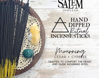 May include: A wooden tray holds a bundle of black incense sticks with yellow tips, alongside crystals and a doily. Text reads "Salem Supply Co." and "Hand Dipped Ritual Incense Sticks". The label says "Mourning" with cedar and cypress.