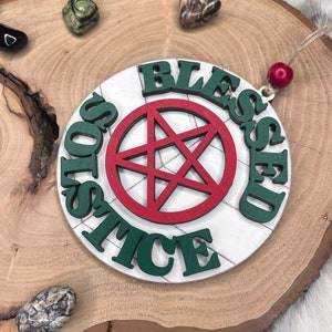 May include: A white wooden ornament with a red pentagram and the words "Blessed Solstice" in green letters.