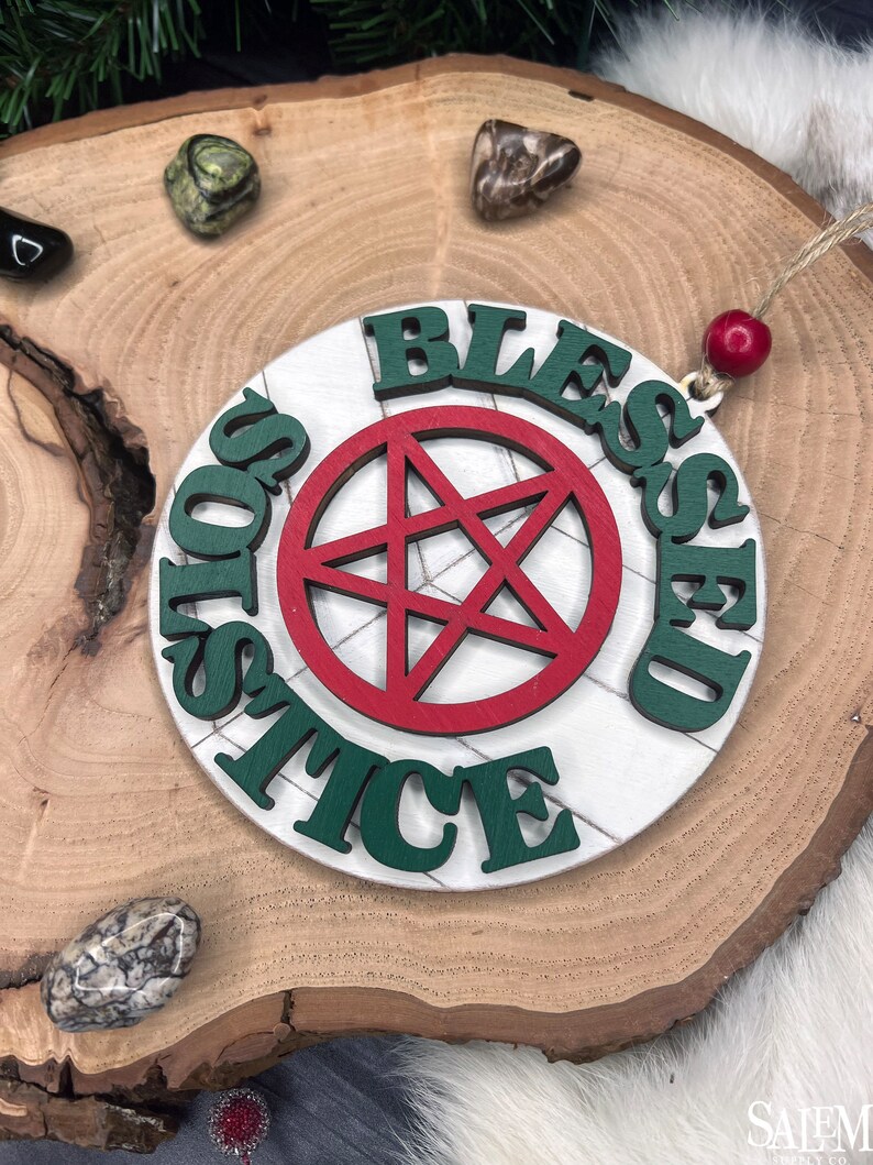 May include: A white wooden ornament with a red pentagram and the words "Blessed Solstice" in green letters.