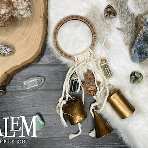 May include: A white macrame dream catcher with a wooden ring inscribed with the text "RING ONE BELL FOR EVIL SPIRITS GET YOU REPEL ALLOW TO PASS THOSE WHO MEAN NO HARM IS IT LET BE." The dream catcher has a brown leather tassel, a gold hamsa charm, and three small glass bottles with dried herbs. The dream catcher is surrounded by various crystals and stones on a wooden surface.