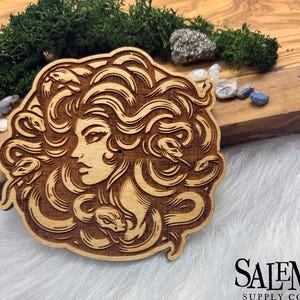 May include: A wooden cutout of a woman with long, flowing hair, surrounded by snakes. The woman's face is visible, and she has a serious expression. The cutout is a light brown color and has a detailed design.