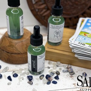 May include: Two small glass bottles containing green liquid with black dropper tops. The bottles are labelled "astral" and have a white background with black text. The bottles are on a wooden surface with a stack of tarot cards and small blue and white stones.