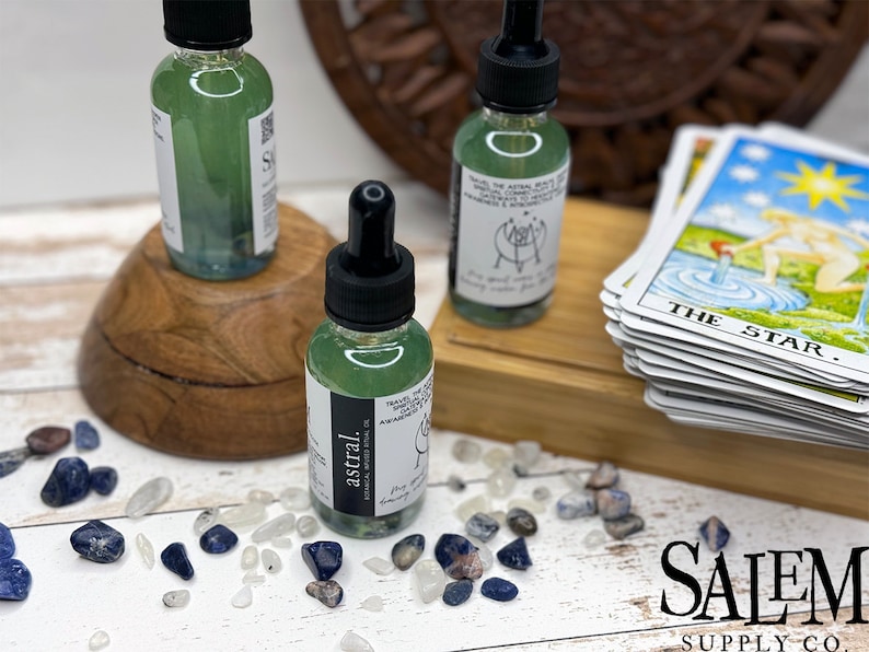 May include: Two small glass bottles containing green liquid with black dropper tops. The bottles are labelled "astral" and have a white background with black text. The bottles are on a wooden surface with a stack of tarot cards and small blue and white stones.