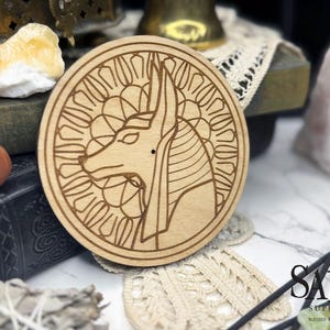 May include: A round, light-colored wooden coaster featuring an engraved image of Anubis, the Egyptian god of the afterlife. The coaster is surrounded by a decorative border. The text "Salem Supply Co." is visible in the lower right corner.