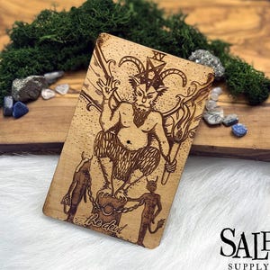 May include: A wooden tarot card with a detailed illustration of a horned demon figure with a pentagram on its head. The card is labeled "XV" and "The Devil".