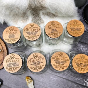 May include: Eight glass jars with wooden lids, each lid featuring a different herb and its associated magical properties. The herbs include Bay Leaf, Star Anise, Wormwood, Mugwort, Hyssop, Red Clover, Nettle, and Vervain.