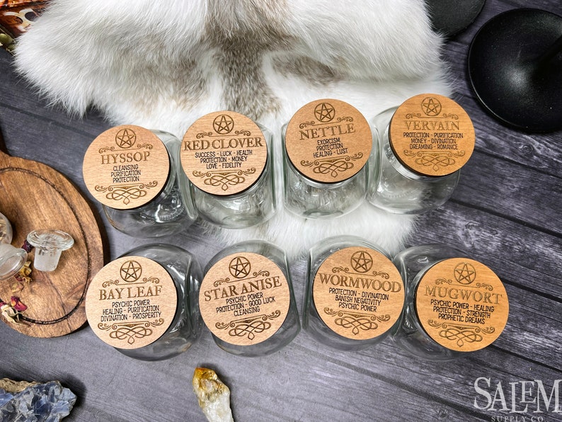 May include: Eight glass jars with wooden lids, each lid featuring a different herb and its associated magical properties. The herbs include Bay Leaf, Star Anise, Wormwood, Mugwort, Hyssop, Red Clover, Nettle, and Vervain.