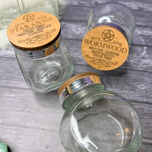 May include: Three glass jars with wooden lids. The lids are engraved with the names of herbs: Star Anise, Wormwood, and another herb. The jars are on a wooden surface.