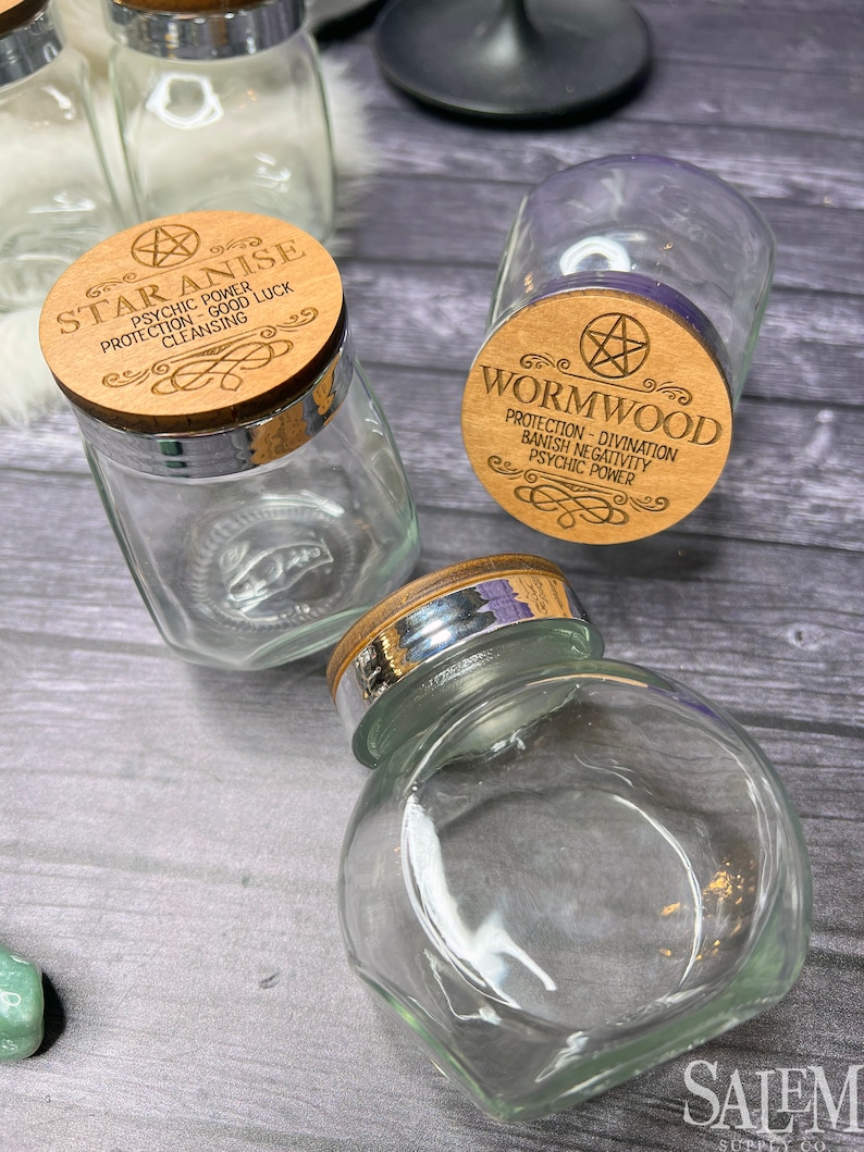 May include: Three glass jars with wooden lids. The lids are engraved with the names of herbs: Star Anise, Wormwood, and another herb. The jars are on a wooden surface.