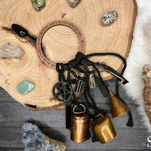 May include: A wooden circle with the inscription "LOVE GIVE BEST SPELLS EVIL SPIRITS THEY DO REPEL ALLOW TO PASS THOSE WHO MEAN NO HARM" and a black pentagram charm hanging from it. There are three gold bells, a small bottle, a key, and black ribbon attached to the circle. The circle is on a wooden surface with various crystals.