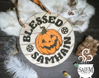 May include: A round wooden sign with the text "Blessed Samhain" in black lettering. A carved pumpkin with a jack-o'-lantern face is in the center of the sign. The sign has a white background with a wood grain pattern.