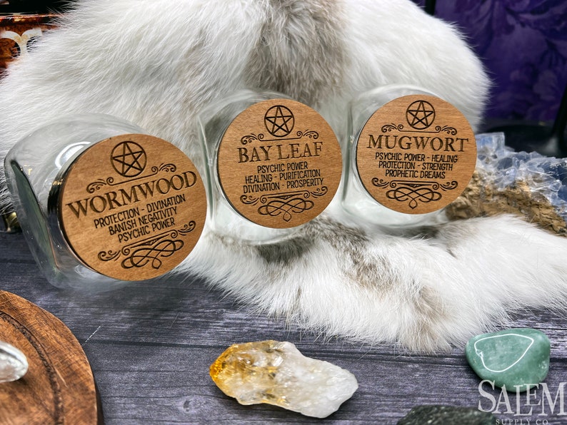 May include: Three glass jars with wooden lids. Each lid has a pentacle symbol and text describing the herb inside. The jars are labeled "Wormwood", "Bay Leaf", and "Mugwort".