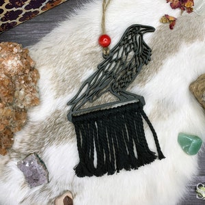 May include: Black wooden crow cutout with a black tassel hanging from the bottom. The crow is perched on a branch and has intricate details. The crow is surrounded by crystals and other decorative items.