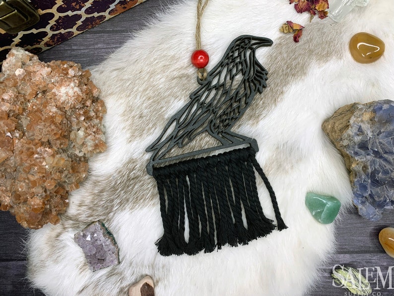 May include: A dark gray wooden raven ornament with a black macrame fringe. The raven is perched on a base, with a red bead above it, suspended by twine. The ornament is displayed on a white fur-like surface.