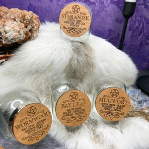 May include: Four glass jars with wooden lids. Each lid has a pentagram and the name of a herb: Star Anise, Wormwood, Bay Leaf, and Mugwort. Each lid also lists the herb's properties, such as psychic power, protection, and healing.