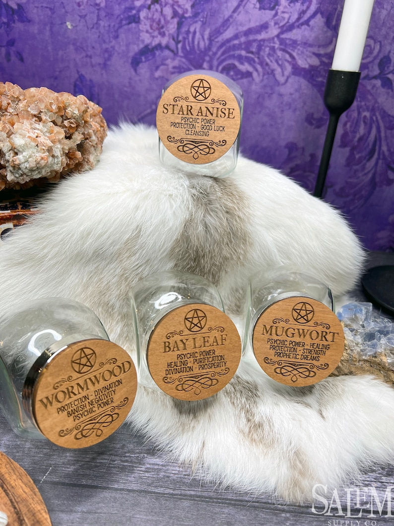 May include: Four glass jars with wooden lids. Each lid has a pentagram and the name of a herb: Star Anise, Wormwood, Bay Leaf, and Mugwort. Each lid also lists the herb's properties, such as psychic power, protection, and healing.