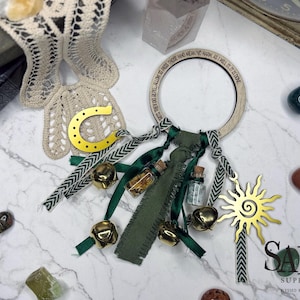 May include: A wooden ring with the text "THEY DO REPEL ALLOW TO PASS THOSE WHO MEAN NO HARM, AS I WILL IT, IT IS DONE." is adorned with green ribbons, small bells, and miniature glass bottles. A gold horseshoe and sun charm are also attached.