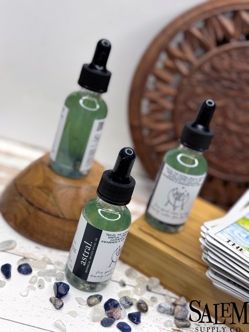 May include: Three glass bottles containing green liquid with black droppers. The bottles are labelled 'astral' and feature text about 'travel the astral' and 'spiritual connection'. The bottles are positioned on a wooden surface surrounded by small blue and white stones.