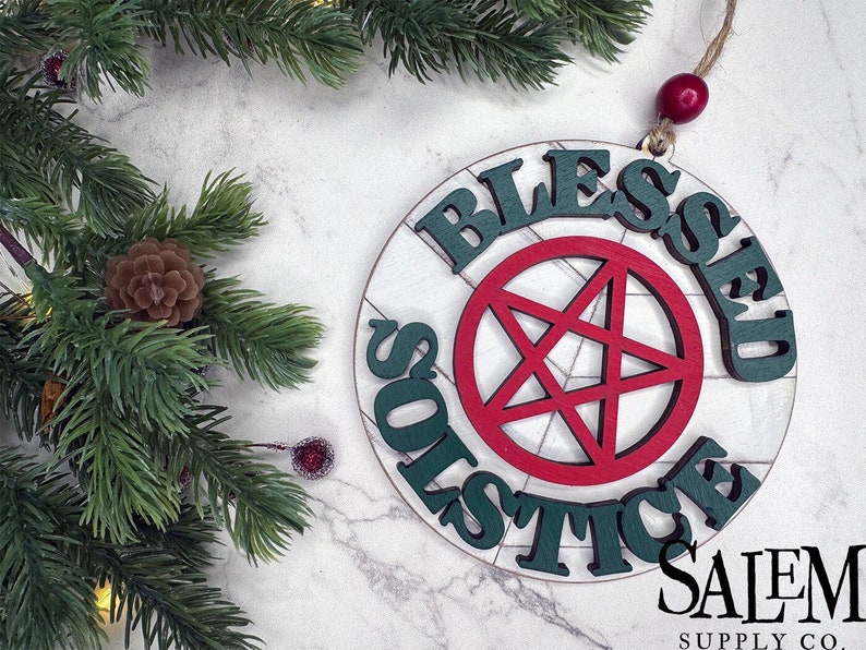 May include: A round wooden ornament with the words 'Blessed Solstice' written in green letters around the edge. In the center of the ornament is a red five-pointed star with a white outline. The ornament is hanging from a string with a red bead at the top. The text 'Salem Supply Co.' is written in white letters on a black background below the ornament.
