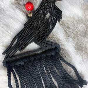 May include: Black raven-shaped wall hanging with a macrame fringe. The raven is made of layered wood and has a detailed cutout design. A red bead hangs from a jute string at the top. The macrame fringe is made of black rope.