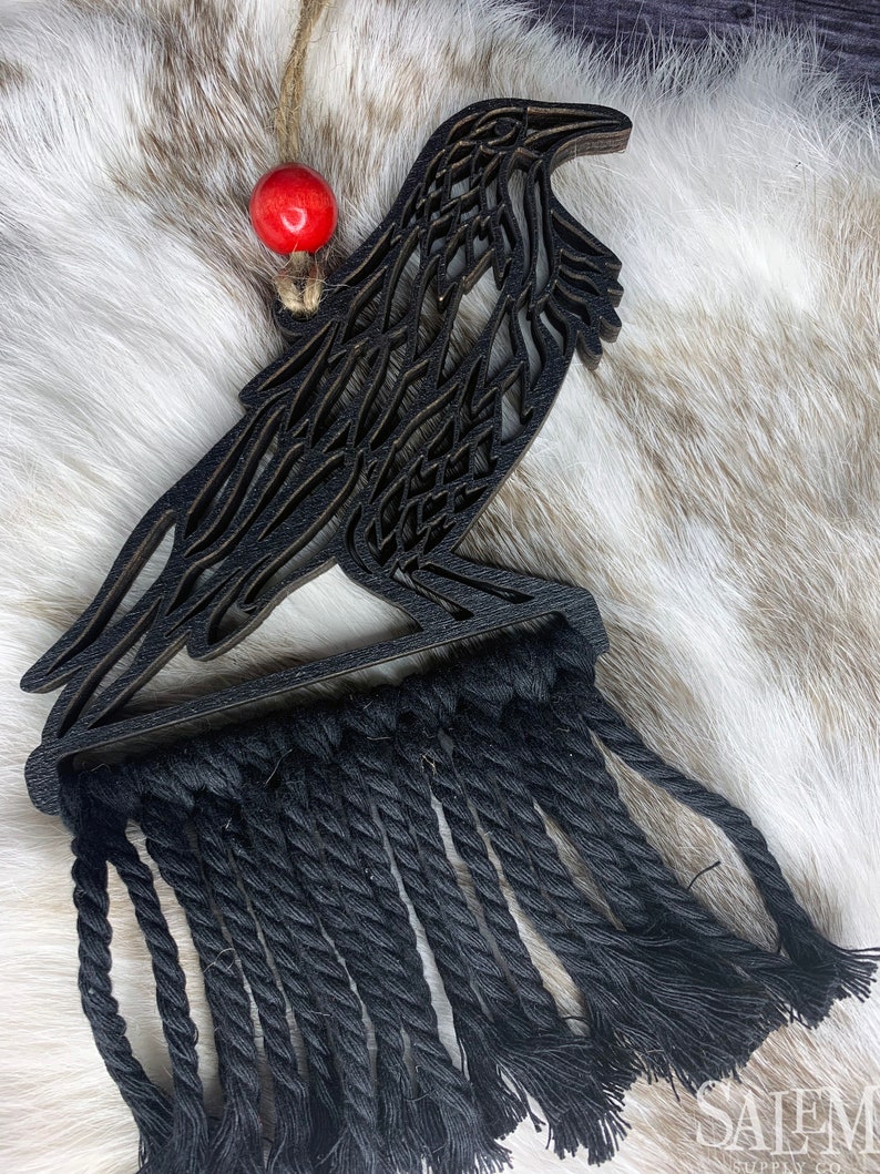 May include: Black raven-shaped wall hanging with a macrame fringe. The raven is made of layered wood and has a detailed cutout design. A red bead hangs from a jute string at the top. The macrame fringe is made of black rope.