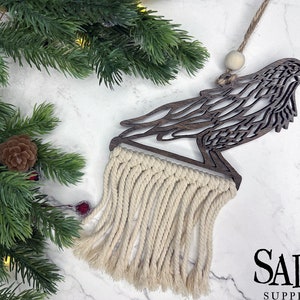 May include: A wooden raven ornament with a macrame fringe. The raven is cut from dark wood and has intricate feather detailing. The macrame fringe is cream-colored. The ornament hangs from a twine string with a wooden bead.