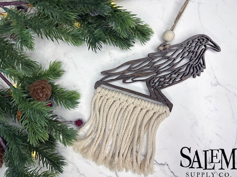 May include: A wooden raven ornament with a macrame fringe. The raven is cut from dark wood and has intricate feather detailing. The macrame fringe is cream-colored. The ornament hangs from a twine string with a wooden bead.