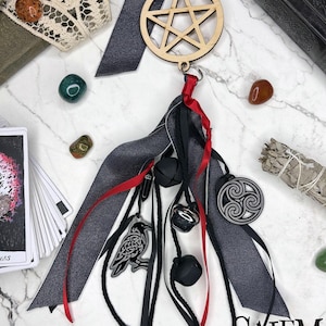 May include: Wooden pentagram charm with black and gray ribbons, red accents, and dangling charms. Charms include a raven, bells, and a Celtic symbol. The image also includes crystals, tarot cards, and a bundle of sage.