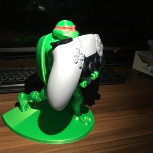 Puede incluir: Una figura verde impresa en 3D de una Tortuga Ninja Mutante Adolescente sosteniendo un mando blanco de PlayStation 5. La figura tiene una máscara roja y está arrodillada sobre una base verde, diseñada para sostener el mando. El mando es blanco con detalles negros.