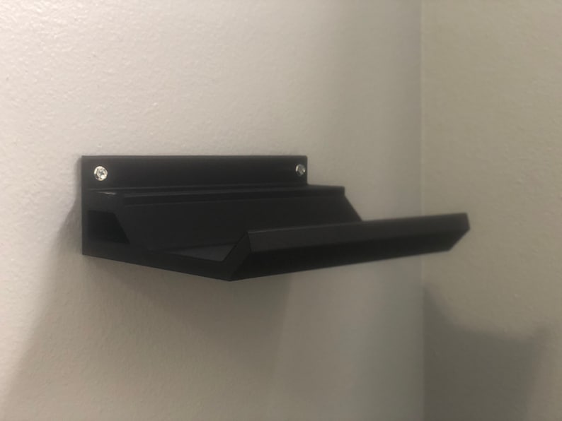 May include: A black wall-mounted shelf with two angled shelves. The shelf is made of plastic and has a simple design.