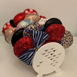 May include: A white stand with a triangular pattern holds a collection of colourful sequined Minnie Mouse ears. The ears are in various colours, including red, blue, gold, pink, and black.