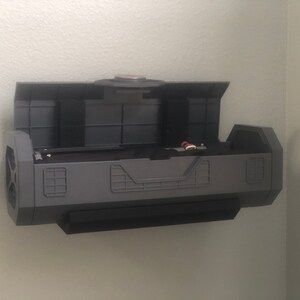 May include: A gray wall-mounted storage unit with a hinged lid open, revealing an empty interior. The unit has a textured, industrial design with a black base.