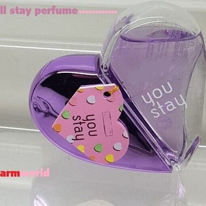 May include: A heart-shaped perfume bottle with a purple metallic base and a clear, curved top. The bottle contains a lavender-colored liquid and has the words "you stay" printed on it. A pink tag with the words "you stay" and colorful polka dots hangs from the bottle.