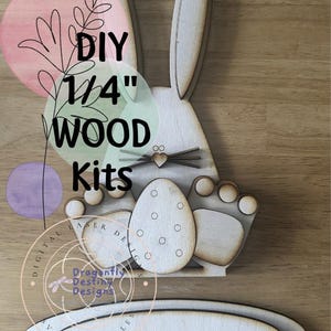 May include: A wooden DIY Easter bunny kit with an egg and paw prints. The text "DIY 1/4" WOOD Kits" is visible. The kit includes a base and is made of light-colored wood.